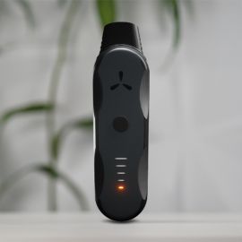 Vaporisateur à herbes AirVape XS Go debout sur une surface blanche avec LED orange allumée, plante floue en arrière-plan.