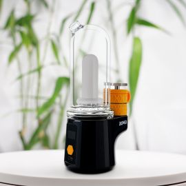 Un Bomb Sykloud E Rig noir élégant avec un récipient en verre et une chambre de chauffe recouverte d'un revêtement orange, présenté sur une surface blanche avec un arrière-plan flou composé de feuilles vertes.